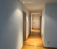 A clean, well-lit hallway with wooden flooring, white walls, and recessed ceiling lights. Doors are visible on both sides, leading to other rooms.