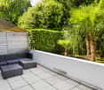 Modern outdoor seating area with wicker sofa, glass table, and ottoman, surrounded by lush greenery and palm trees.