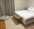 A large bed, white sheets, white mattress, pillows, a bench, rug, and a curtain in front of the bed.