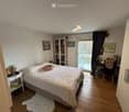 Wooden floor, white walls, bed with white bedding, bedside table with figurines, glass door leading to a balcony, a bookshelf with books and figurines, framed photos on the wall, and a desk by the window. A lamp is mounted on the ceiling. The room has a c