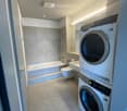 Modern bathroom with a bathtub, toilet, sink, and stacked washing machine/dryer unit. The space features light-colored tiles and a sleek design.