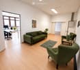 Modern living room with light wooden flooring, green sofa, armchairs, small table, lamp, and a plant. Large windows provide natural light, and a small office area is visible through an open doorway.