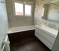 Modern bathroom with a bathtub, sink, and mirror. Features dark tiled flooring and a window with frosted glass.