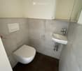 Modern bathroom with a toilet, sink, and mirror. The walls are tiled in light grey, and the floor is dark brown.