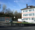 A multi-story residential building with white walls, green shutters, and a brown roof. The building has multiple windows and a chimney. There is a fenced area in front with a parked car, a van, and a basketball hoop.