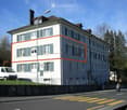 A large, multi-story residential building with white walls, green shutters, and multiple windows. The building has a classic architectural style with a pitched roof and chimney. A white van is parked in front, and the building is situated near a road with