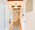 A narrow hallway with wooden flooring, white walls, and a striped runner. Features a hanging light fixture and several doors leading to other rooms.