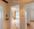 Bright, modern living space with wooden flooring, white walls, and large windows. Features a sofa, small table, chairs, and a hanging lamp.
