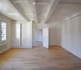 Empty hallway with wooden flooring, white walls, and several closed doors leading to other rooms. A window on the left side allows natural light to enter.