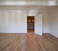 Empty room with wooden flooring and white walls, featuring an open doorway leading to a small kitchen area.