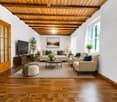 Modern living room with wooden floors, exposed wooden ceiling beams, large windows, and a mix of neutral and blue tones. Features a sofa, coffee table, ottoman, armchair, side tables, lamps, and a television.