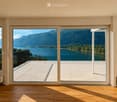 Bright, empty room with wooden flooring and large sliding glass doors opening to a balcony overlooking a scenic lake and mountains.