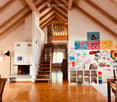 Bright, open living space with wooden floors, exposed wooden beams, fireplace, staircase leading to a loft area, and colorful children's artwork displayed on the walls.