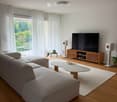Modern living room with a grey sectional sofa, a white coffee table, a large flat-screen TV on a wooden stand, and two white speakers. The room features wooden flooring, large windows with white curtains, and a balcony with outdoor furniture visible throu
