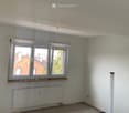 Empty room with white walls, large window with wooden sill, radiator, and visible electrical outlets. No furniture or personal items present.