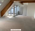 Empty attic room with wooden beams, large windows, and carpeted floor.