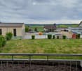 Balcony overlooking a grassy area with a fence, several buildings, parked cars, and piles of sand and gravel in the background.