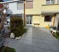 Patio area with tiled floor, small table, chairs, and greenery near a two-story house.