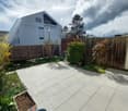 Garden area with tiled patio, wooden fence, various plants, and a modern house in the background.