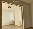 Empty room with light-colored tiled flooring, white walls, and a large window allowing natural light. Features a radiator and a ceiling light fixture.