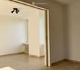 Empty room with beige tiled flooring, white walls, and a large open doorway leading to another space. A small window and radiator are visible in the adjacent area.