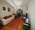 Living room with a cream-colored sofa, red and brown throw pillows, a coffee table, a bookshelf, a table lamp, a television, a vase, a chair, and a plant. The room features wooden flooring and white walls.