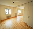 Empty room with wooden flooring, white walls, and two windows with radiators underneath. The room is connected to another space through an open doorway.