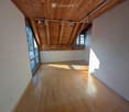 Empty room with wooden flooring, white brick walls, and exposed wooden beams on the ceiling. Natural light streams in through large windows, casting shadows on the floor.