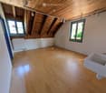 Empty attic room with wooden floors, exposed wooden beams, white walls, and two windows. A white sink is visible in the corner.