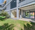 Modern residential building with a well-maintained garden area, featuring a lawn, small trees, and a paved patio with large glass doors leading to the interior.