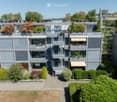Modern multi-story residential building with grey facade, balconies, and greenery. Features include multiple levels, outdoor terraces, and surrounding landscaped gardens.