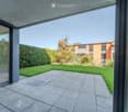 Modern outdoor patio with large glass doors leading to a well-maintained lawn and surrounding greenery, with a contemporary building in the background.