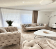 Modern living room with large windows and balcony access, featuring a beige tufted sofa, coffee table, ottoman, floor lamp, TV stand, and a potted plant.