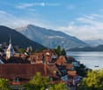 A picturesque town with traditional red-roofed houses, a prominent church tower, and a serene lake surrounded by lush green hills and distant mountains.