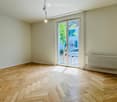 Empty room with wooden herringbone flooring, white walls, large glass sliding door leading outside, radiator on one wall, and a single hanging light fixture.