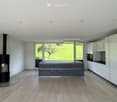 Modern kitchen with white cabinetry, dark grey island, built-in oven, and a black wood-burning stove. Large windows provide natural light and a view of the greenery outside.