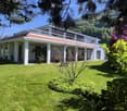 A well-maintained lawn with a modern two-story house featuring a red-tiled roof and white columns, surrounded by lush greenery and flowering trees.