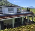 Two-story house with a flat roof terrace, surrounded by greenery and mountains in the background.