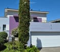 Modern two-story house with white and purple exterior, large garage, and well-maintained landscaping including a tall evergreen tree and shrubs.