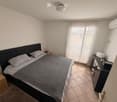 A bed with grey pillows and a blanket, nightstands on either side, wooden cabinet with drawers, air conditioner, white curtains on the balcony