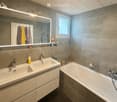 Modern bathroom with double sink, bathtub, large mirror with LED lighting, and various toiletries on the counter.