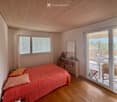 Bright bedroom with wooden floors, a large bed with orange bedding, a nightstand, a bookshelf, and a sliding glass door leading to a balcony with outdoor furniture.