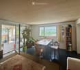 Bright, open room with wooden ceiling, large windows, and sliding glass doors leading to a patio. Features a sofa, dining table with chairs, bookshelf, and a large potted plant.