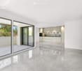 Open-plan space with large glass sliding doors leading to an outdoor area, white tiled floor, and a modern kitchen area with white cabinets and stainless steel appliances.