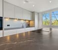 Modern kitchen with white cabinets, dark marble countertops, stainless steel sink, and built-in oven. Large windows provide natural light.