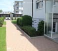 Well-maintained garden area with neatly trimmed bushes and a paved walkway leading to a modern building entrance.