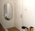 Clean, minimalist hallway with white walls, tiled floor, and a shoe rack holding multiple pairs of shoes. A round mirror on the wall reflects part of the adjacent room.