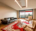 Modern living room with a slanted ceiling, large windows, and a balcony. Features a black leather sofa, white armchairs, a coffee table, and a red patterned rug.