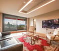 Modern living room with large windows offering a scenic view, featuring a leather sofa, coffee table, armchairs, and a television.