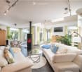 Modern living room with white sofas, colorful cushions, a glass coffee table, a central fireplace, large windows with yellow curtains, and a dining area with chairs and a table.